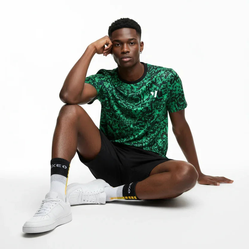 A male model sits on a white studio floor wearing a green patterned athletic t-shirt, black shorts, white socks, and white sneakers.