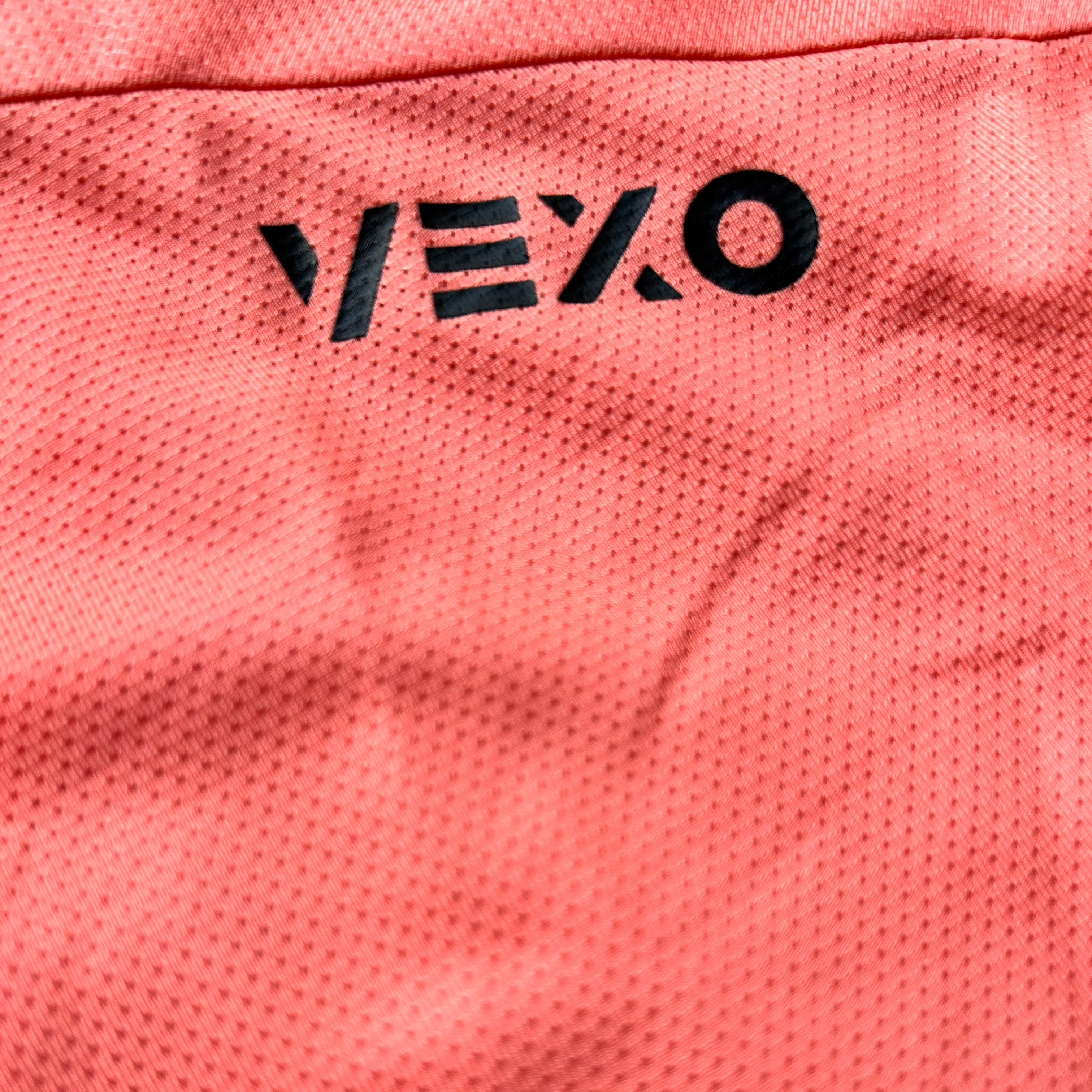 Close-up of a coral pink perforated athletic fabric with a black vertical Vexo logo printed on the side.