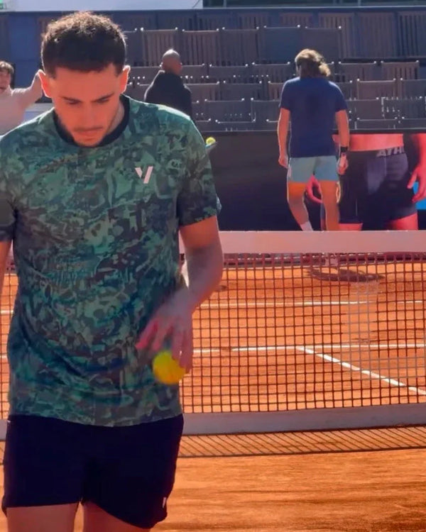 Tennis player in a green patterned shirt serving a yellow ball on a clay court.