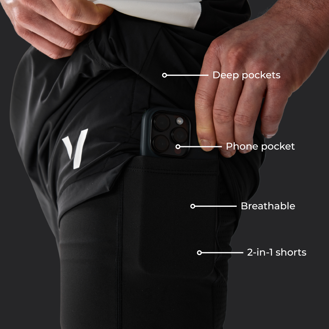 Close-up of a person wearing black athletic shorts with labeled callouts pointing to deep pockets, phone pocket, breathable fabric, and 2-in-1 shorts. The person is pulling up the shorts, and a smartphone is shown inside the side pocket.