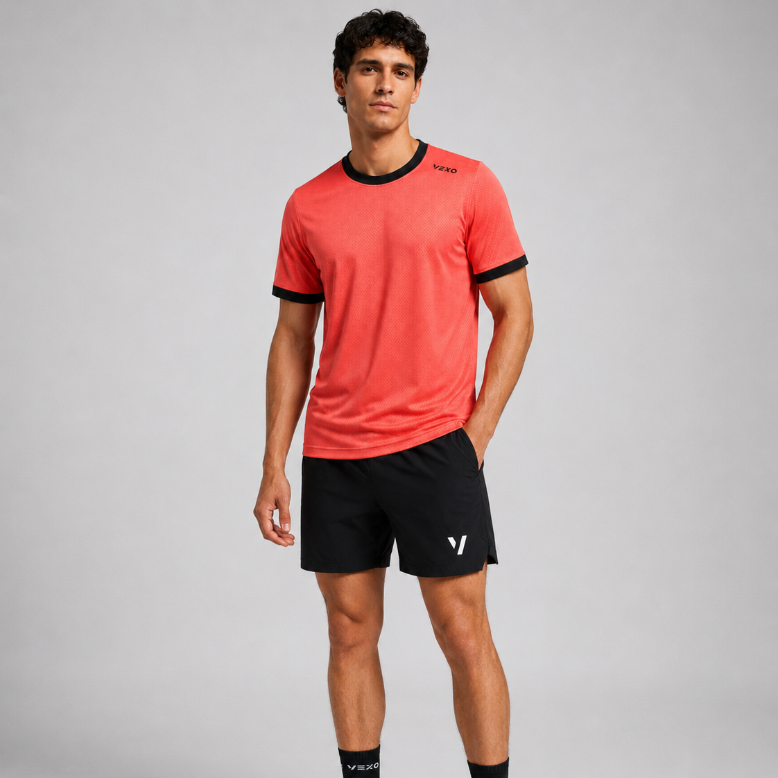 A man modeling a coral red athletic t-shirt with black trim and black athletic shorts, paired with white sneakers and black socks, against a light gray studio background.
