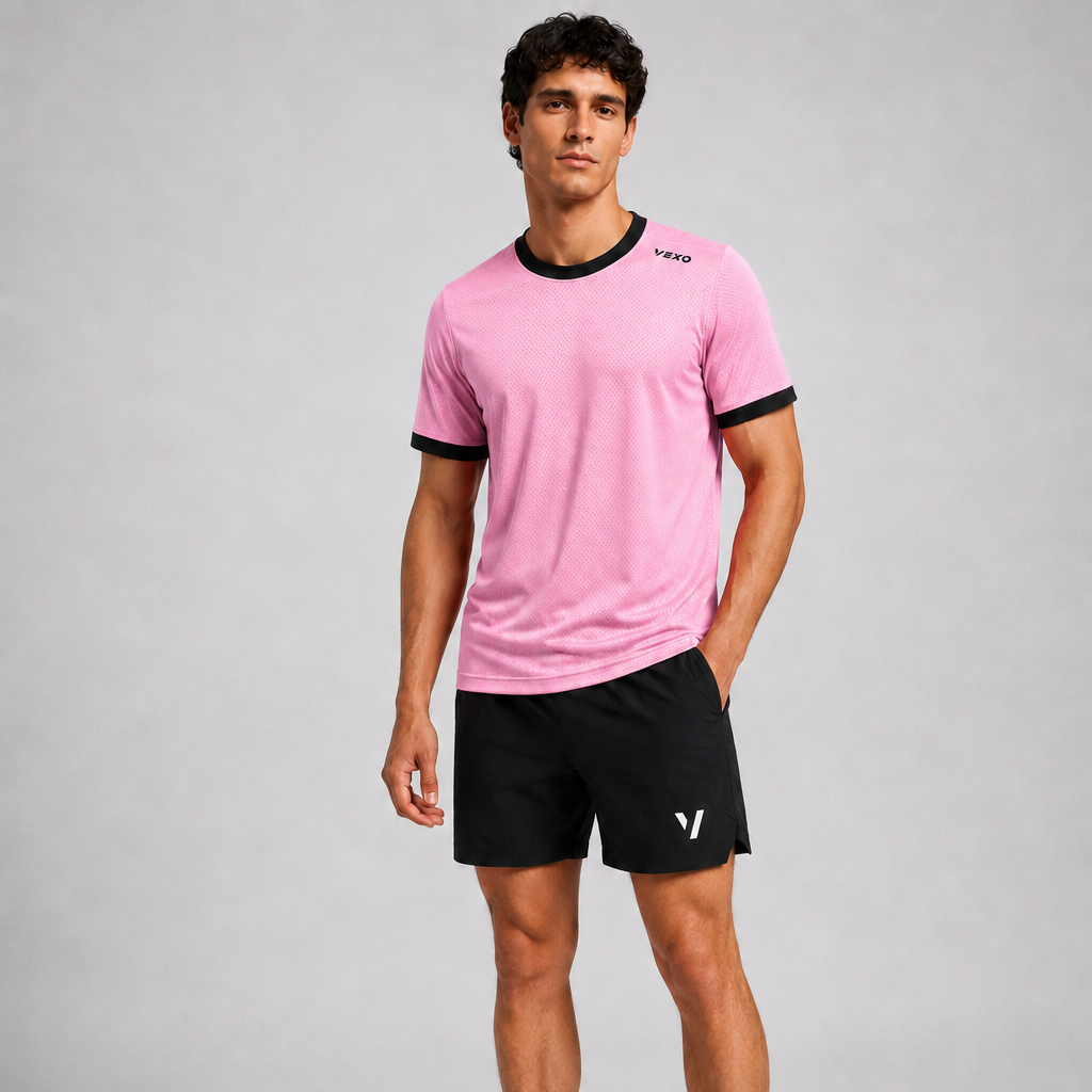 A man modeling a pink short-sleeve athletic t-shirt with black trim, black athletic shorts, white socks, and white sneakers against a light gray studio background.